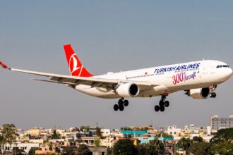 A white Turkish Airlines aircraft is positioned to the centre right of the image, shown in flight. The nose of the aircraft is close to the right edge of the image, the tail close of the centre. The body of the aircraft has Turkish Airlines branding along the side towards the front. The tail of the aircraft is red. The background shows a blue sky and rooftops of buildings.