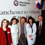 Three women and two men stood in front of a sign that reads "Manchester to Orlando", for Manchester Airport.
