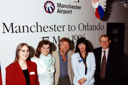 Three women and two men stood in front of a sign that reads "Manchester to Orlando", for Manchester Airport.
