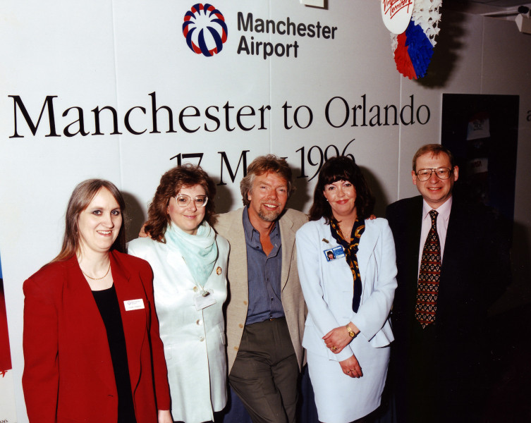 Three women and two men stood in front of a sign that reads "Manchester to Orlando", for Manchester Airport.