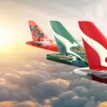 Tails of Qantas group aircraft- Qantas and Jetstar, mid-flight. Clouds beneath them, the sun in the background.