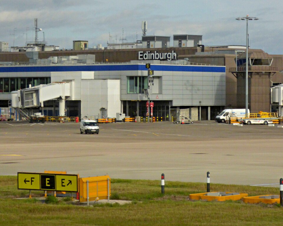 Edinburgh Airport airside area showing terminal buildings, boarding gates, ground vehicles, and taxiway signage.