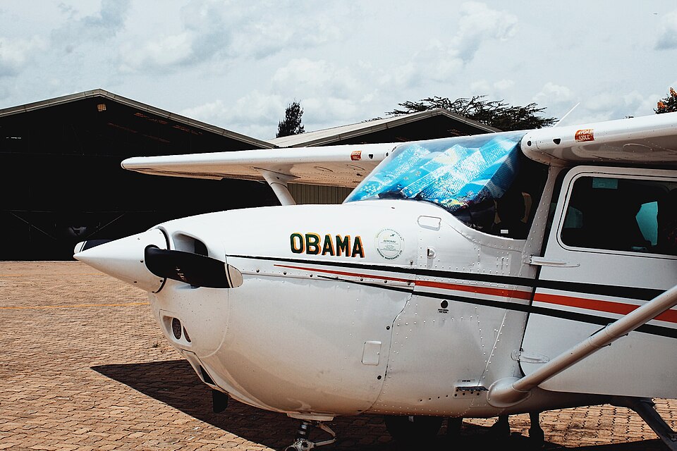 A close-up, side-angle photograph of the front fuselage and cockpit area of a white Cessna light utility airplane. The aircraft features horizontal red and black racing stripes along its body.