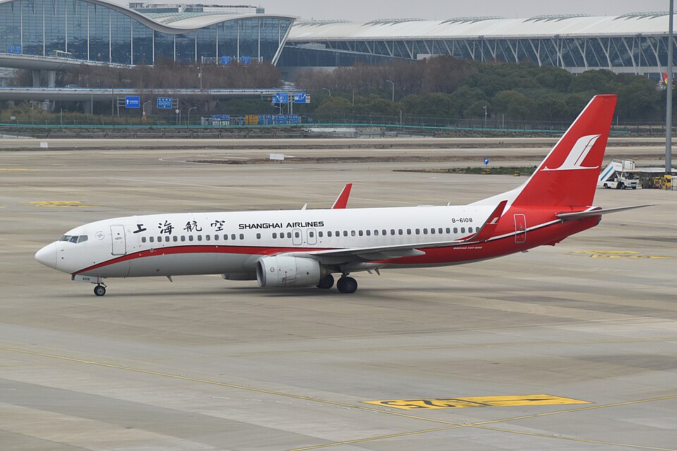 A red-and-white Shanghai Airlines Boeing 737-800 taxis across the apron at Shanghai Pudong International Airport, with the terminal building visible in the background.