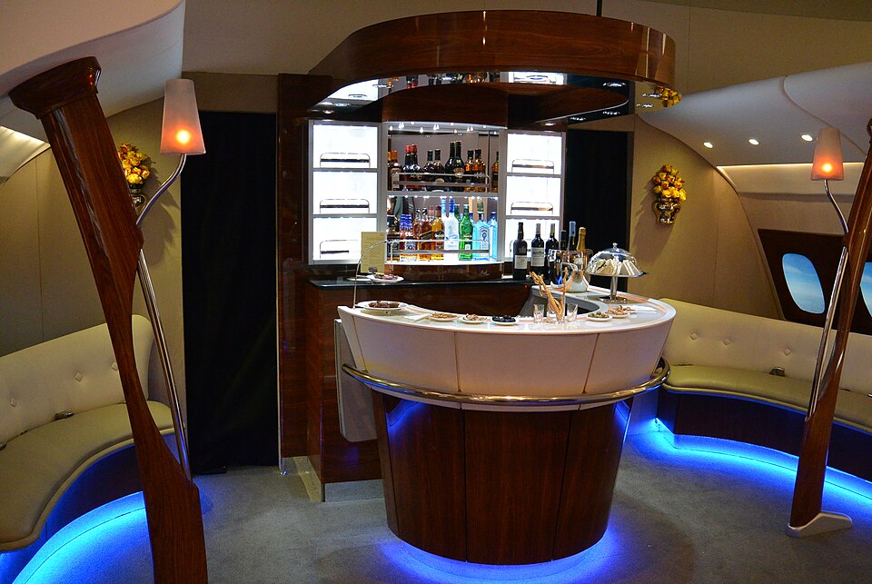 Interior of an Emirates Airbus A380 onboard lounge featuring a curved bar counter with drinks, snacks, and glassware, with seating and illuminated shelves in the background.