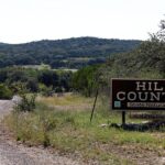 A rural landscape with a dirt road running through grassy terrain and trees, with low, forested hills in the background.