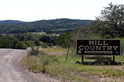 A rural landscape with a dirt road running through grassy terrain and trees, with low, forested hills in the background.