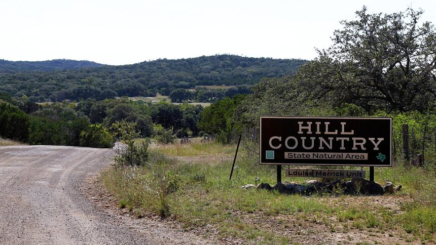 A rural landscape with a dirt road running through grassy terrain and trees, with low, forested hills in the background.