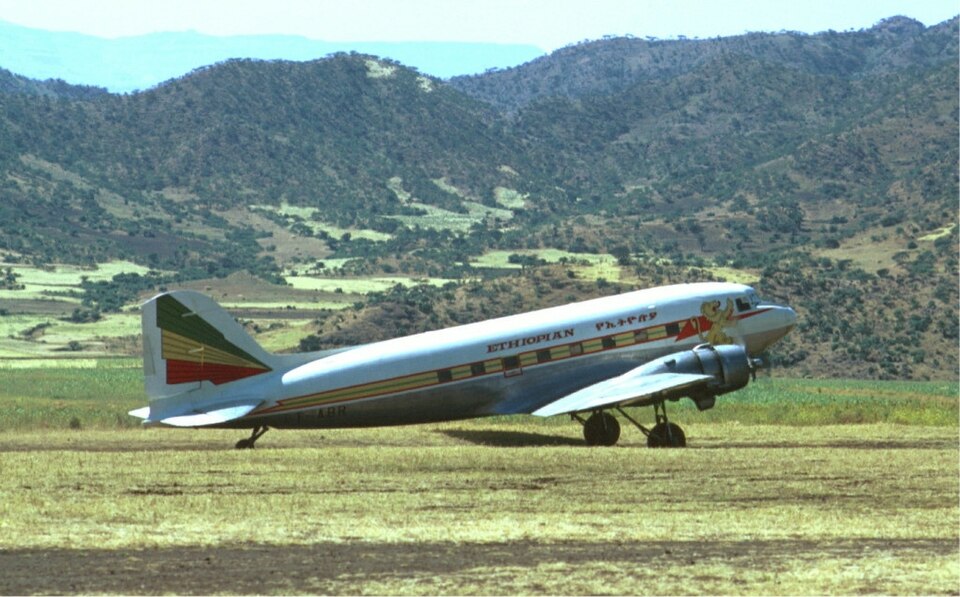 Vintage photo of an Ethiopian Airline aircraft from 1974. The plane is seen taking off in front of a green and mountainous landscape. The plane has a lion on its side and is mostly white.