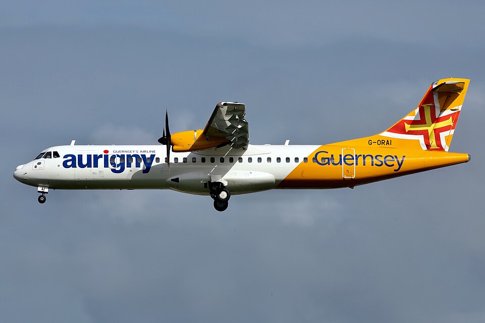 Aurigny aircraft flying in the air. Grey-ish sky in the background. The aircraft has landing gear out, and has white and yellow livery. Aurigny is written in dark blue, with a yellow and red cross on the tail.