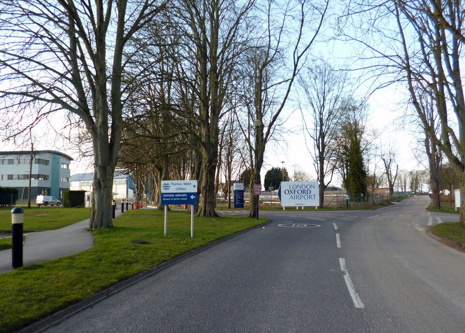 Entrance road to London Oxford Airport lined with tall trees, with airport signage and nearby buildings visible.
