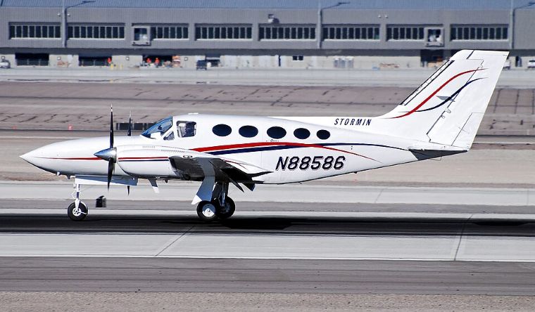 A light twin-engine aircraft on a runway at an airport, with visible registration markings and terminal buildings behind it. 