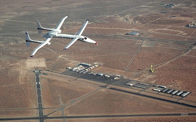 An aerial view of the Scaled Composites Proteus research aircraft soaring over Las Cruces International Airport in New Mexico, with desert terrain and intersecting runways visible below.