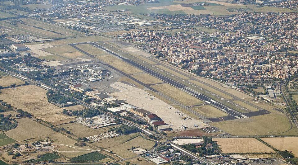 Aerial view of Rome Ciampino Airport showing a long runway, parked aircraft, airport buildings, surrounding suburbs, and farmland under clear skies.