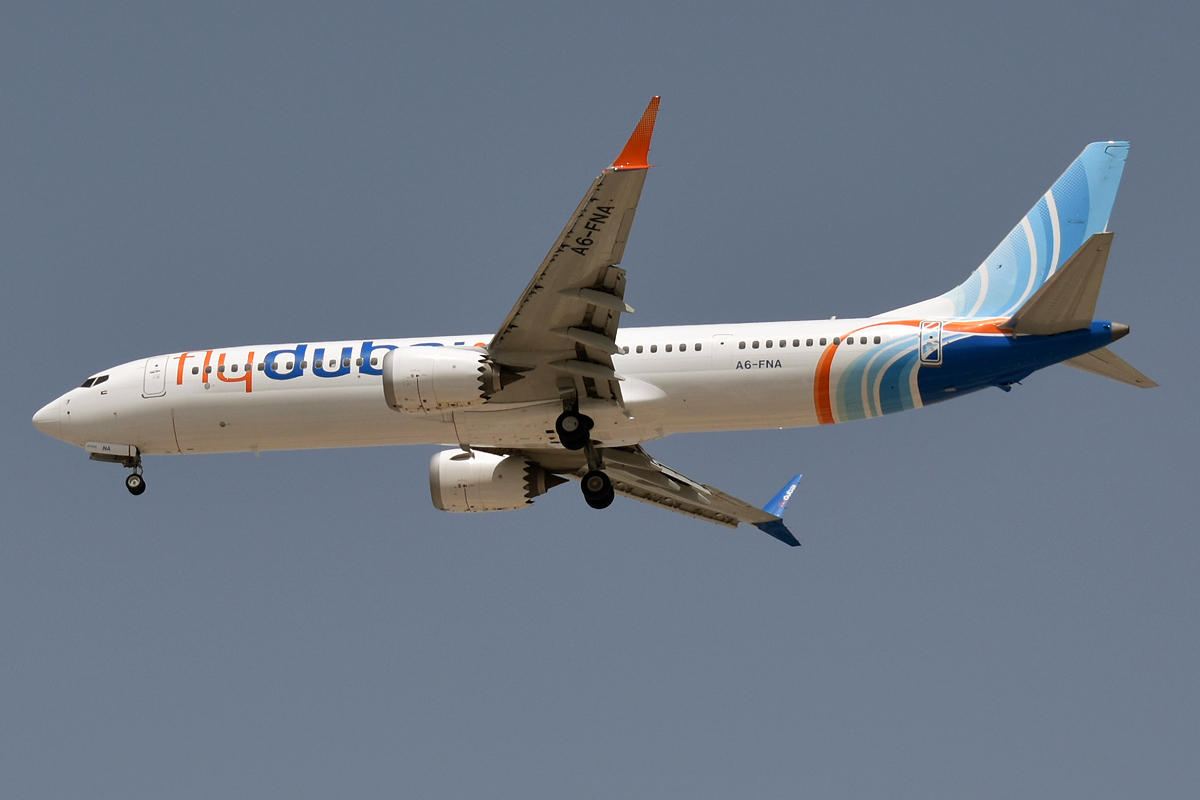 A B737 Flydubai aircraft flying under a grey sky. The aircraft's tail is light blue with white stripes and an orange stripe.