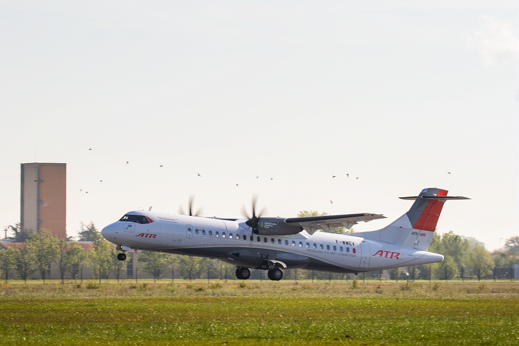 An ATR72-600 aircraft in ATR house livery, registration F-WNEV, lifting off from a runway with propellers in motion, trees and a tower visible in the background.