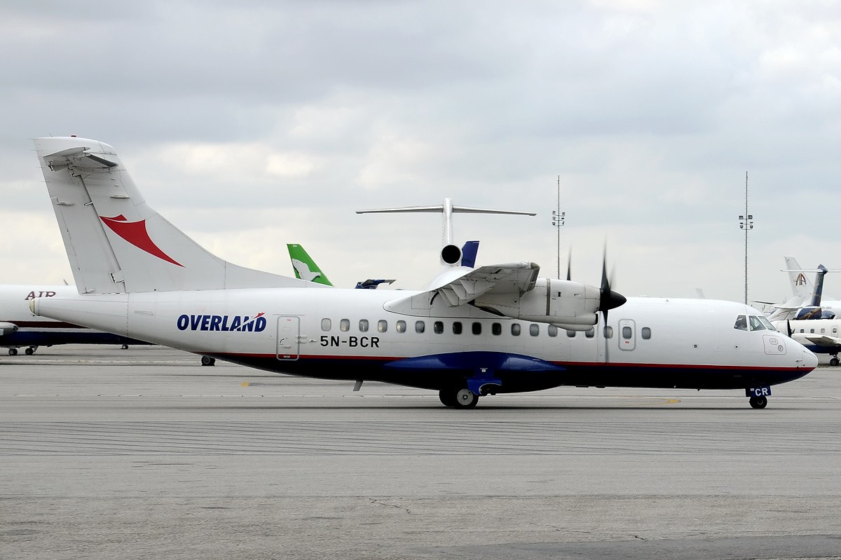 An ATR-42-320 model Overland Airways aircraft is pictured from the side at an airport taxiway. the nose is positioned centre right of the image and the tail is positioned centre left. The plane is mostly white with a dark blue underbelly. It has propellers, and a T -shaped tail, which has a red Overland Airwards logo on it. The lower back end of the plane has blue and red 'Overland' branding. 