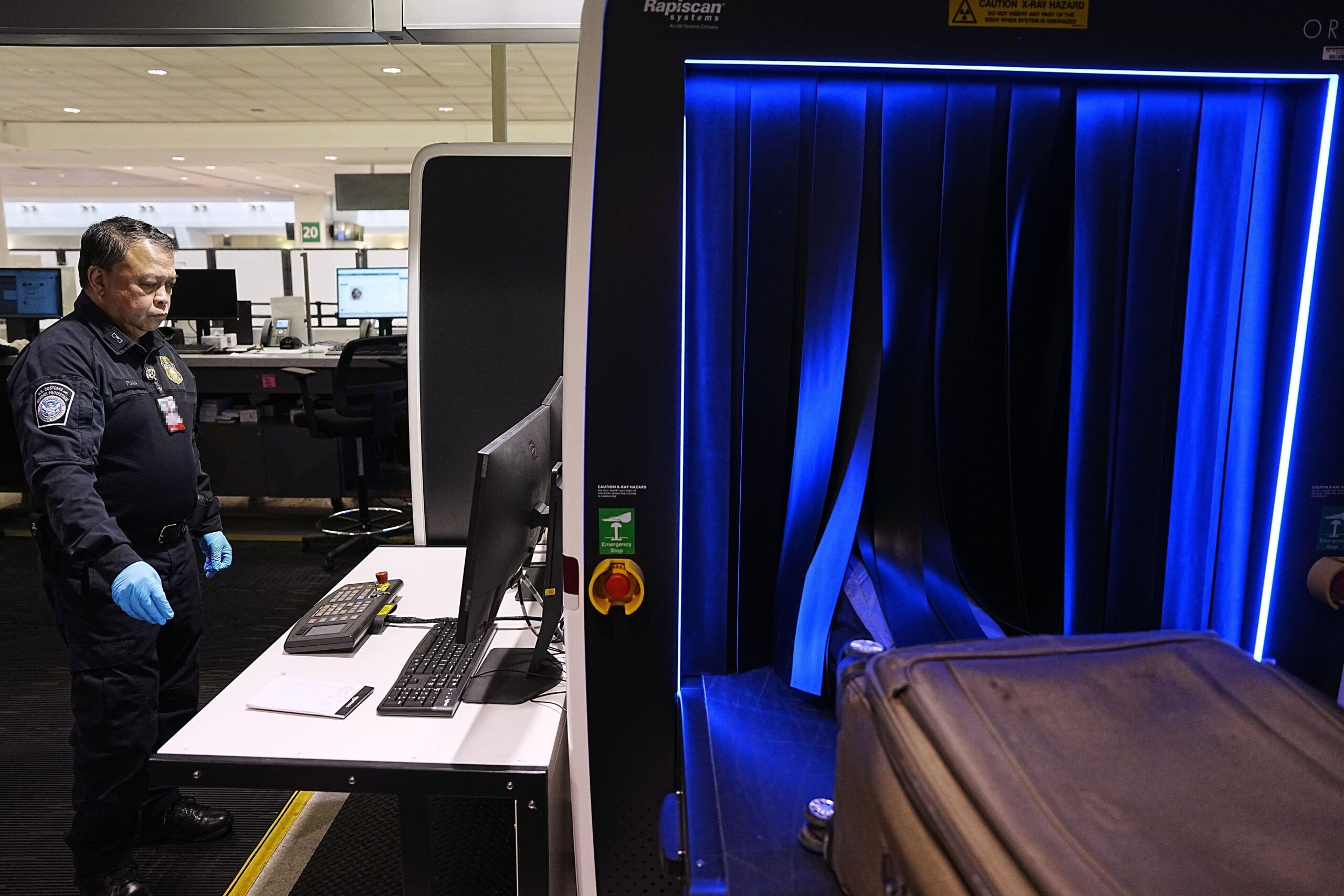 Interior view of an airport security screening area, with an X‑ray baggage scanner in the foreground illuminated by blue lighting around the conveyor opening. A suitcase sits on the moving belt entering the machine through dark rubber flaps. To the left, a uniformed security officer wearing blue protective gloves stands at a workstation with a computer monitor, keyboard and control panel. Behind the officer, an office‑like space with desks, chairs and additional screens is visible under bright overhead lighting, while warning labels about X‑ray hazards are displayed on the scanning equipment.