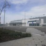 Wide view of a modern, low‑rise airport terminal building set back from a quiet access road. The building is white and grey with long horizontal windows and glass entrance doors, topped with rows of rooftop ventilation units. In the foreground, a pedestrian crossing with bold white stripes leads across the empty road, bordered by paved walkways, low shrubs and newly planted trees supported by wooden stakes. Tall streetlights line the roadside, and blue pedestrian crossing signs stand near the kerb. The scene appears calm and largely deserted, with no vehicles or people visible, under a pale blue sky lightly veiled with thin cloud.