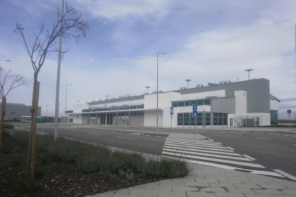 Wide view of a modern, low‑rise airport terminal building set back from a quiet access road. The building is white and grey with long horizontal windows and glass entrance doors, topped with rows of rooftop ventilation units. In the foreground, a pedestrian crossing with bold white stripes leads across the empty road, bordered by paved walkways, low shrubs and newly planted trees supported by wooden stakes. Tall streetlights line the roadside, and blue pedestrian crossing signs stand near the kerb. The scene appears calm and largely deserted, with no vehicles or people visible, under a pale blue sky lightly veiled with thin cloud.
