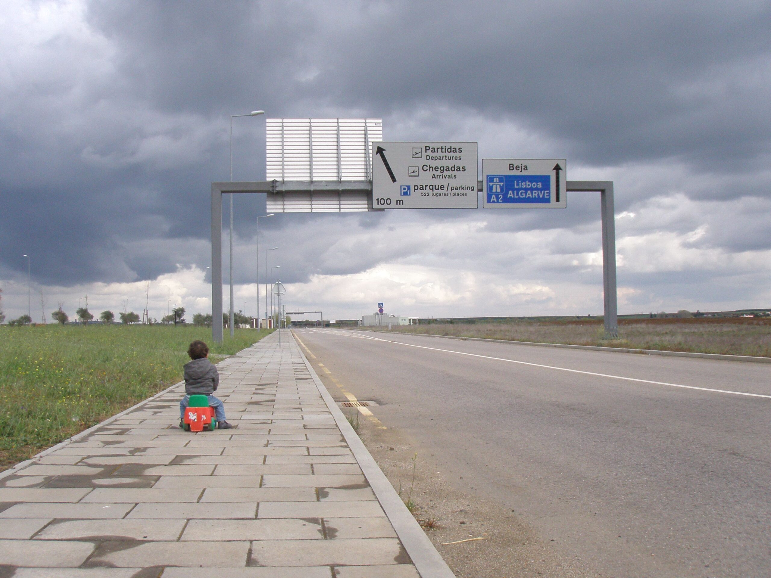 Wide view of a quiet access road leading towards an airport, bordered by a paved pedestrian path on the left and open grassy fields on both sides. In the foreground, a small figure sits on the pavement facing away from the camera, next to a bright red wheeled bag. Overhead road signs span the carriageway, with Portuguese text pointing to Partidas and Chegadas, parking in 100 metres, and directions to Beja, Lisboa and the A2 Algarve. The road stretches into the distance under heavy, dark grey clouds, creating a stark, windswept atmosphere with minimal traffic and flat, open surroundings.
