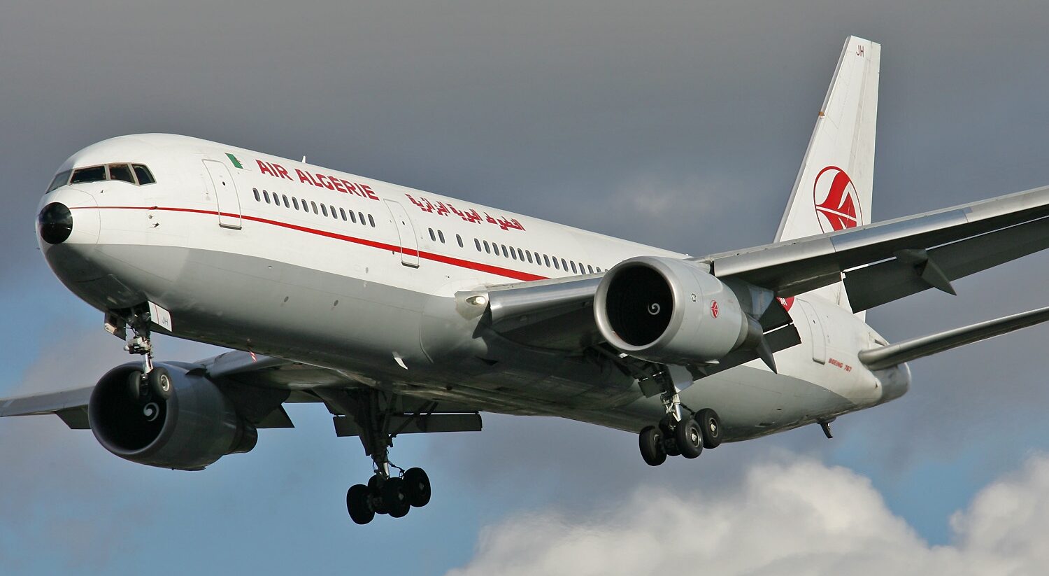 Air Algerie Boeing aircraft flying towards the left side. White and red livery, with a red symbol on the tail. Blue sky in the background with clouds.