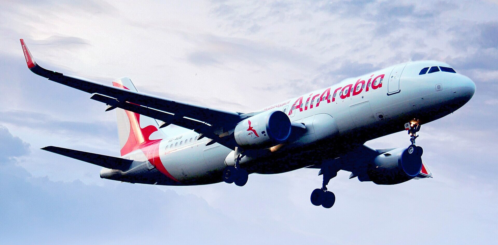 Air Arabia Airbus A320 aircraft flying towards the right side. Blue sky in the background, clouds in the sky. White and red livery.