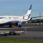 A Boeing 737 Air Peace aircraft is positioned centre left of the image, with the nose positioned right over to the left and the tail in the centre. The aircraft is white with the red blue and white logo on the back end. The sky in the background is blue, and the middle background shows green grass and the foreground shows tarmac.