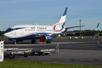 A Boeing 737 Air Peace aircraft is positioned centre left of the image, with the nose positioned right over to the left and the tail in the centre. The aircraft is white with the red blue and white logo on the back end. The sky in the background is blue, and the middle background shows green grass and the foreground shows tarmac.