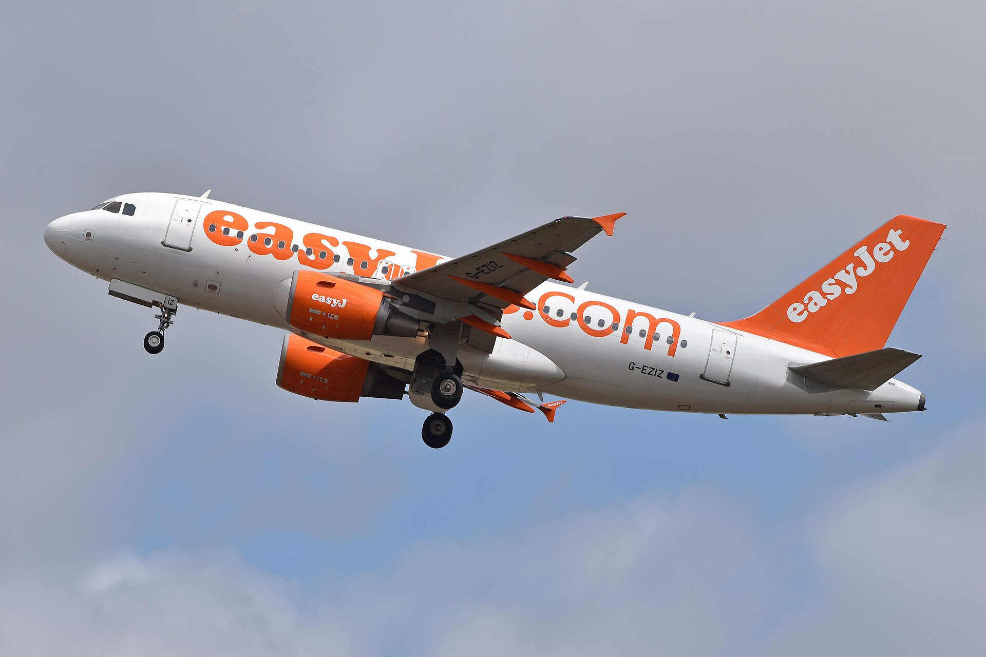 An easyJet aircraft flying towards the left side of the frame. White and orange livery. Blue sky and clouds in the background.