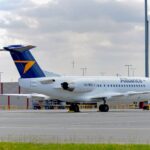 Side view of an Alliance Airlines Fokker 70 parked on an airport apron, with a white fuselage displaying the word “Alliance” in grey lettering and the registration VH‑NKU near the rear. The aircraft has a dark blue tail featuring a bold yellow arrow‑shaped logo, and two rear‑mounted engines integrated into the fuselage. The landing gear is down with wheel chocks in place, and ground handling equipment sits nearby behind low yellow safety railings. In the foreground, a strip of green grass borders the concrete surface, while low airport buildings, light poles and distant hills appear under a cloudy sky.