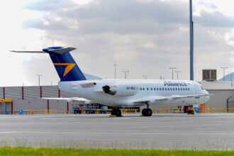 Side view of an Alliance Airlines Fokker 70 parked on an airport apron, with a white fuselage displaying the word “Alliance” in grey lettering and the registration VH‑NKU near the rear. The aircraft has a dark blue tail featuring a bold yellow arrow‑shaped logo, and two rear‑mounted engines integrated into the fuselage. The landing gear is down with wheel chocks in place, and ground handling equipment sits nearby behind low yellow safety railings. In the foreground, a strip of green grass borders the concrete surface, while low airport buildings, light poles and distant hills appear under a cloudy sky.