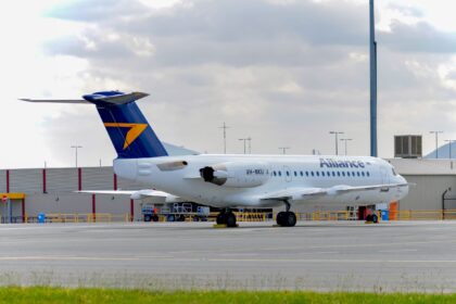 Side view of an Alliance Airlines Fokker 70 parked on an airport apron, with a white fuselage displaying the word “Alliance” in grey lettering and the registration VH‑NKU near the rear. The aircraft has a dark blue tail featuring a bold yellow arrow‑shaped logo, and two rear‑mounted engines integrated into the fuselage. The landing gear is down with wheel chocks in place, and ground handling equipment sits nearby behind low yellow safety railings. In the foreground, a strip of green grass borders the concrete surface, while low airport buildings, light poles and distant hills appear under a cloudy sky.
