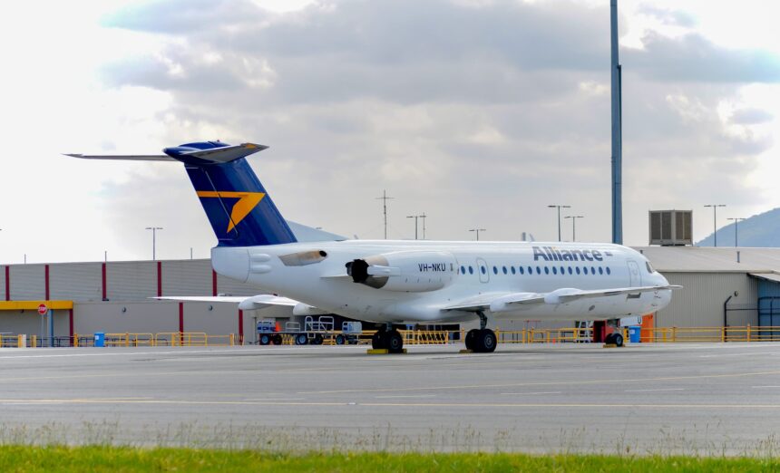 Side view of an Alliance Airlines Fokker 70 parked on an airport apron, with a white fuselage displaying the word “Alliance” in grey lettering and the registration VH‑NKU near the rear. The aircraft has a dark blue tail featuring a bold yellow arrow‑shaped logo, and two rear‑mounted engines integrated into the fuselage. The landing gear is down with wheel chocks in place, and ground handling equipment sits nearby behind low yellow safety railings. In the foreground, a strip of green grass borders the concrete surface, while low airport buildings, light poles and distant hills appear under a cloudy sky.