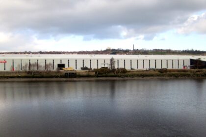 Large BAE Systems industrial building beside a river under cloudy skies, with the company logo visible on the exterior.