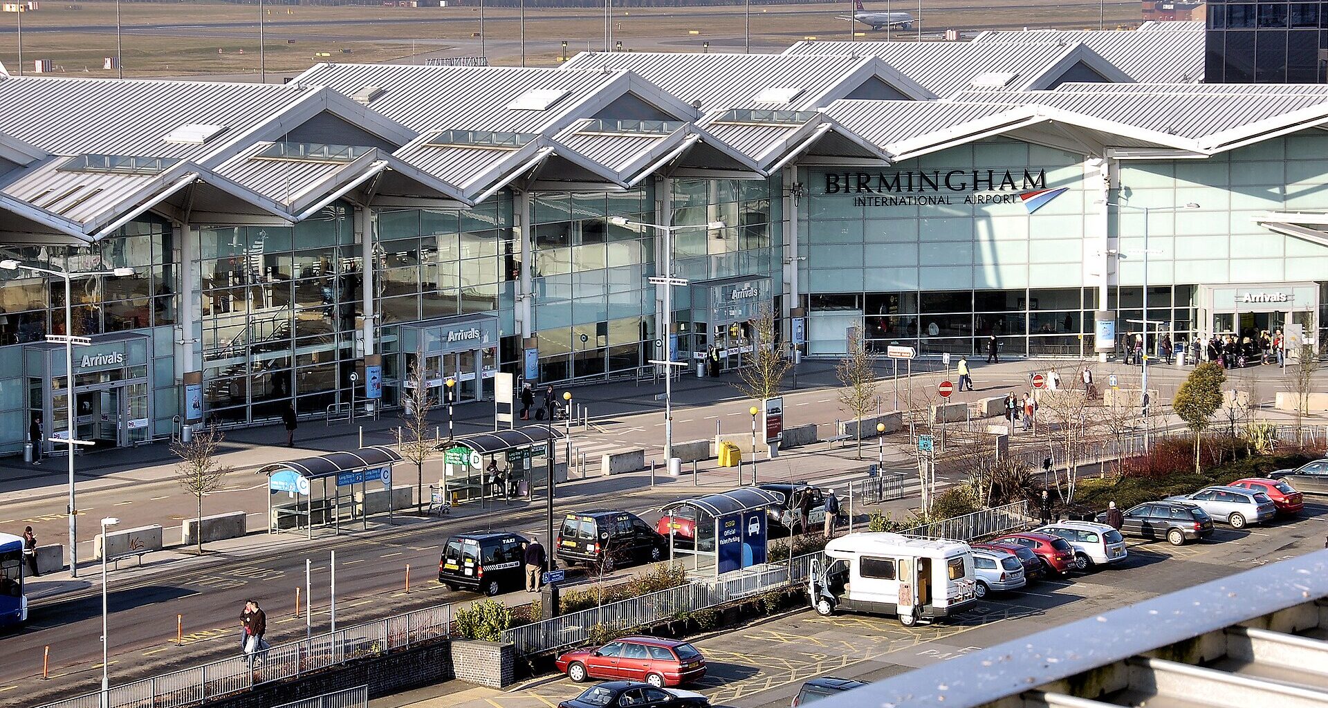 Birmingham airport Terminal 2 exterior. Car park in the foreground. Tall glass windows on the building, with pointed roofs. 