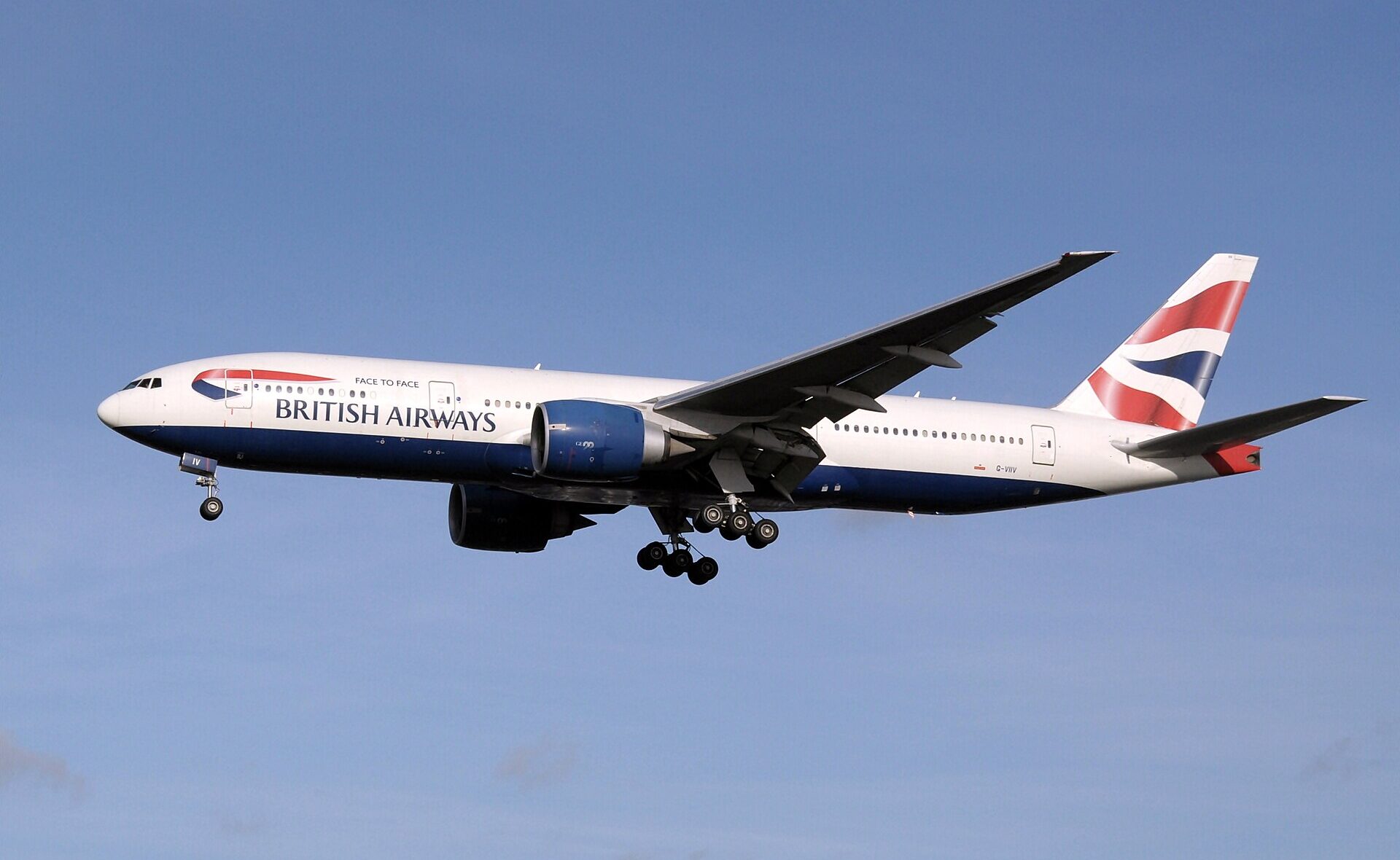 British Airways Boeing 777 flying through the blue sky, with no clouds in the background. The landing gear is out, and it has red, white and blue livery.