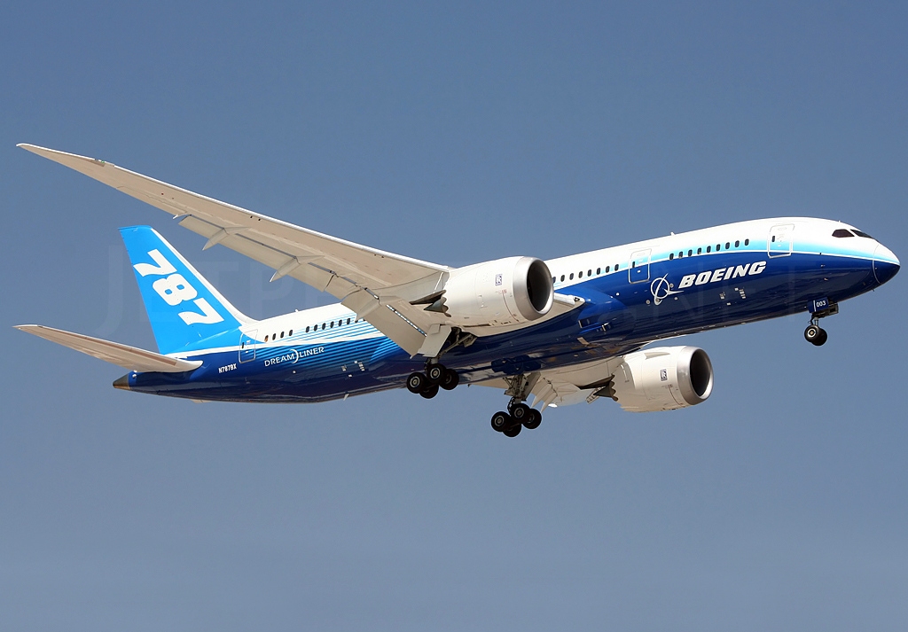 A Boeing 787 aircraft occupied he centre of the image in flight. It is mostly white on top, with a dark blue underbelly and light blue stripe across the middle, and has a light blue tail. White 'Boeing' branding and '787' can be seen on the underbelly and tail. The nose is facing to the upper right centre and the tail is facing to the lower left centre, showing the aircraft at an angle. The background is a clear blue sky.