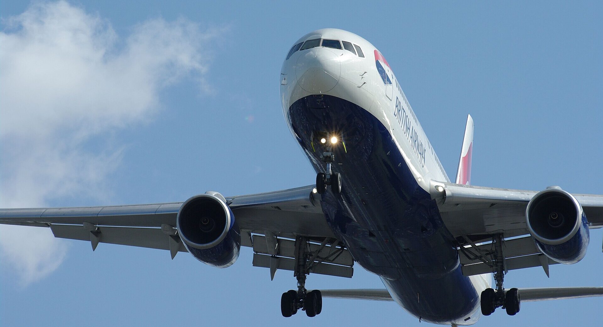 Low-angle shot of a British Airways aircraft flying overhead. Blue sky with clouds in the background. The aircraft has its landing gear out.