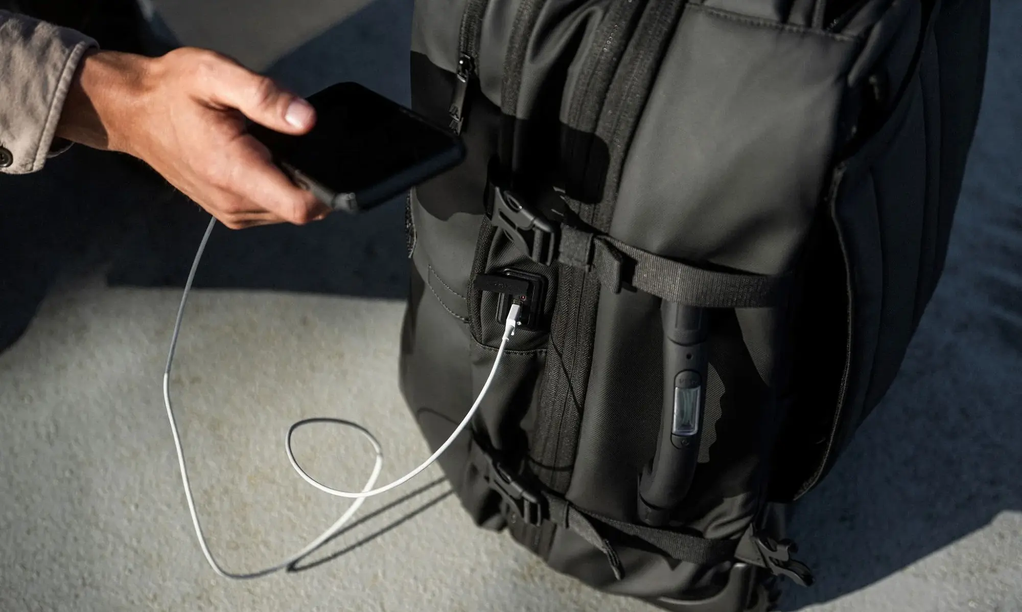 A black backpack on the floor, with a white charging cable coming out from the side, charging a black phone that a man is holding up.