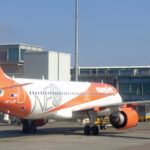 An easyJet aircraft parked at Manchester Airport. White and orange livery. Buildings and blue sky in the background.