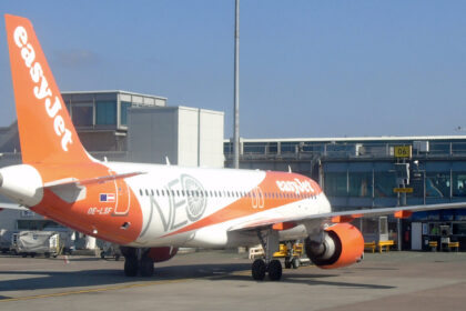 An easyJet aircraft parked at Manchester Airport. White and orange livery. Buildings and blue sky in the background.