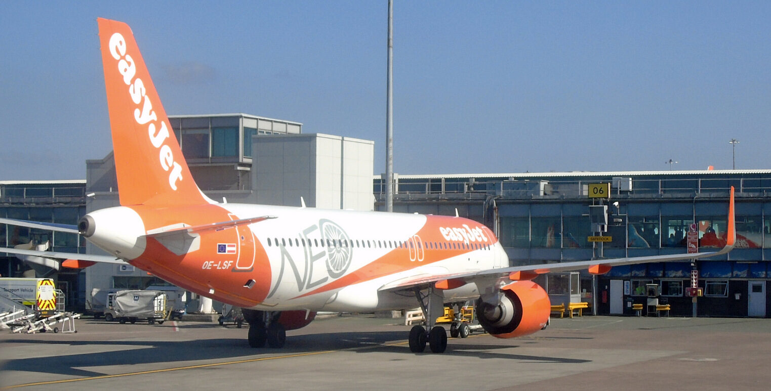 An easyJet aircraft parked at Manchester Airport. White and orange livery. Buildings and blue sky in the background.