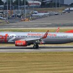 Jet2 aircraft on the Birmingham runway before takeoff. Grass in the foreground, buildings in the background. Grey, red and white livery.