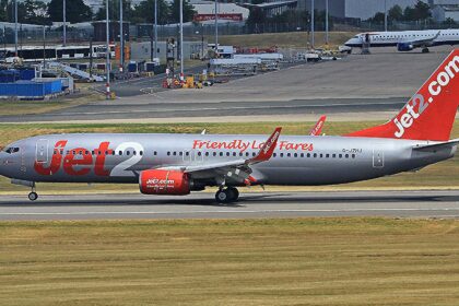 Jet2 aircraft on the Birmingham runway before takeoff. Grass in the foreground, buildings in the background. Grey, red and white livery.