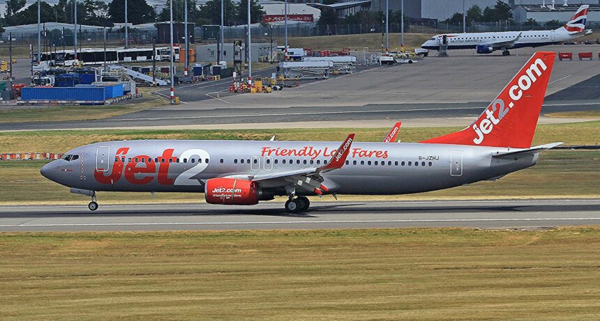 Jet2 aircraft on the Birmingham runway before takeoff. Grass in the foreground, buildings in the background. Grey, red and white livery.