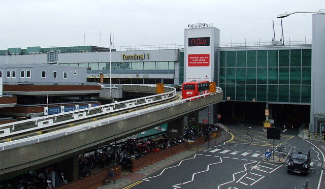 The entrance to London Heathrow Airport Terminal 1 from the top of a building opposite. The road leading up to the car park occupies the bottom right of the image, and the higher bus access road can be seen winding up from the bottom left of the image to the centre. Across the upper centre fro left to right of the image the building can be seen, with the large sign that reads 'Terminal 1' across the top in yellow. The background is a cloudy sky.