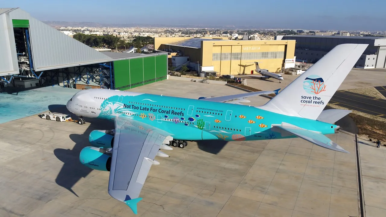 High, angled view of a large Airbus A380 parked on an airport apron outside an aircraft hangar complex. The aircraft is painted in a vivid turquoise and white special livery themed around ocean conservation, with illustrated coral, fish and waves along the fuselage and the slogan “Not Too Late For Coral Reefs” clearly visible on the side. The white tail carries a circular logo with coral imagery and the words “save the coral reefs.” A white pushback tug is connected to the nose landing gear, and the aircraft’s four engines and broad wings dominate the foreground. In the background, large maintenance hangars with green and yellow panels stand alongside smaller business jets, with a sprawling cityscape and low hills visible in the distance under clear blue skies.