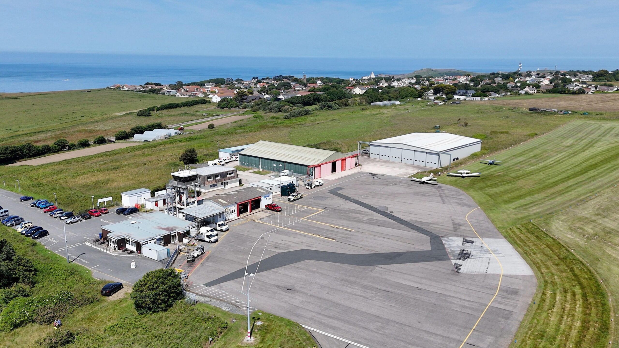 Alderney Airport, on a sunny day taken from above, the coast can be seen in the backdrop in addition to fields and houses.