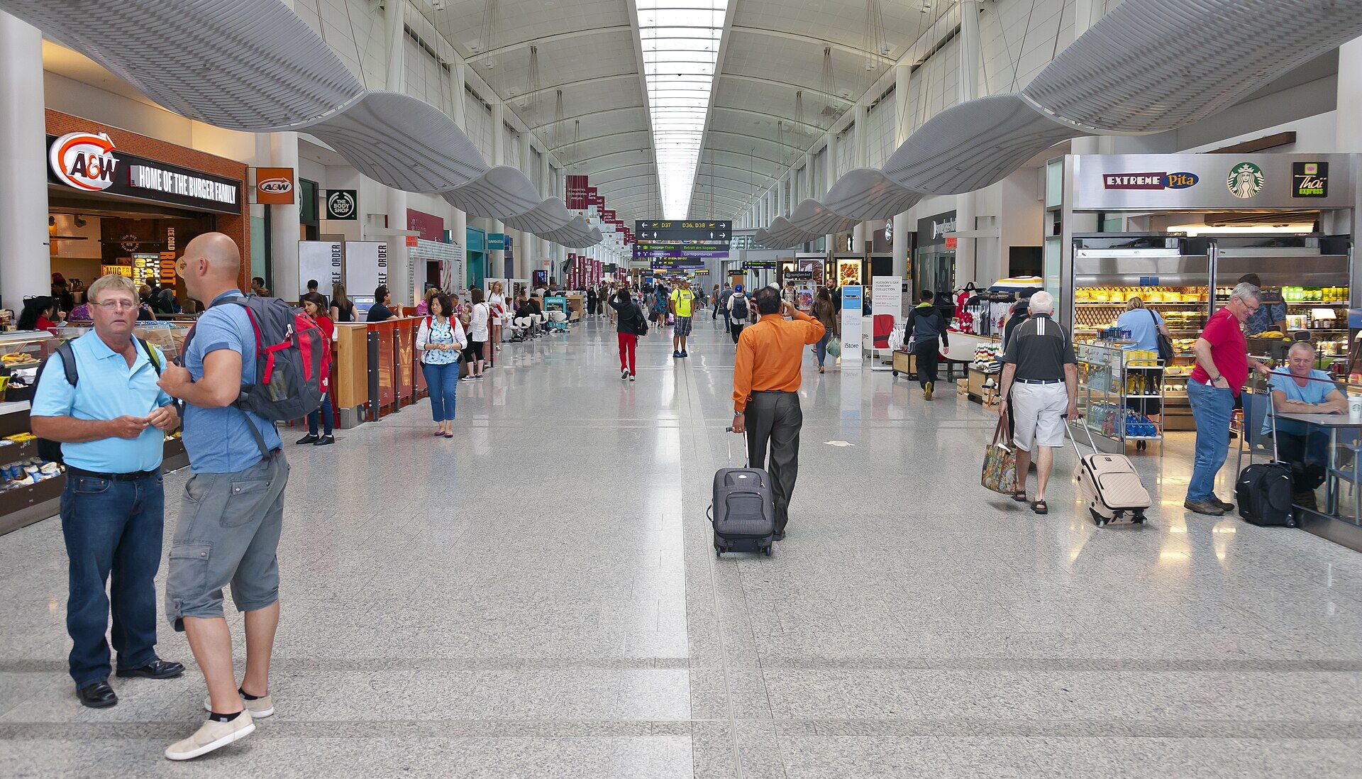 Interior view of Toronto Pearson International Airport. Shops seen both sides of the frame, with travellers with suitcases walking around.